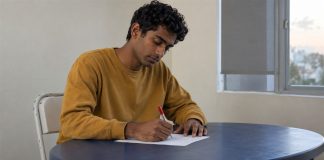A young man writing on a loose sheet of paper at a blue table beside a window