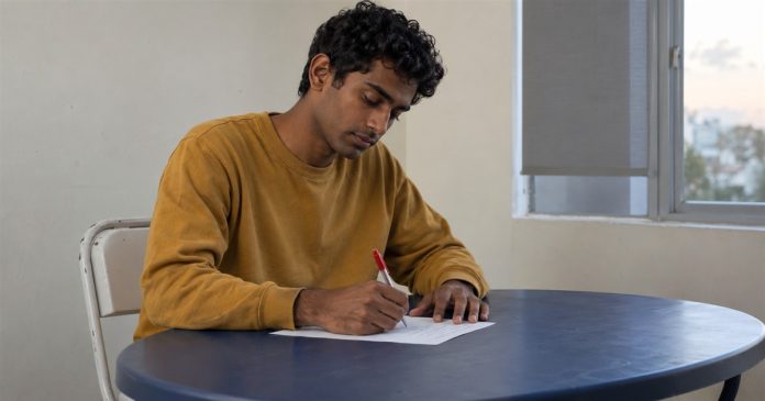 A young man writing on a loose sheet of paper at a blue table beside a window