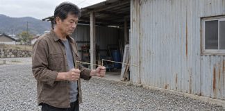 A man holding two brass L-shaped dowsing rods in a gravel courtyard beside a metal shed
