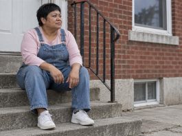 A woman in denim overalls taking a quiet pause on concrete steps outside a brick home