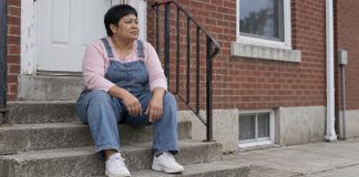 A woman in denim overalls taking a quiet pause on concrete steps outside a brick home