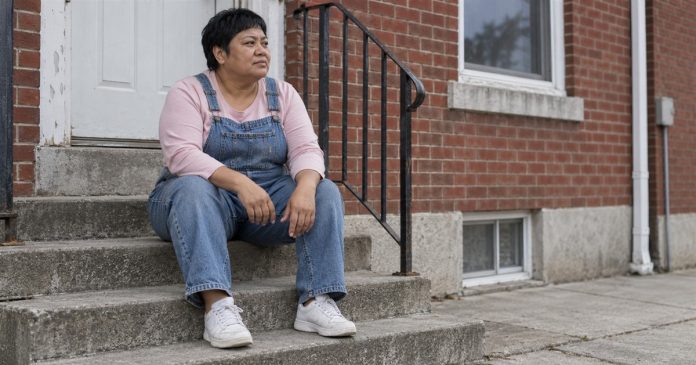 4028 realistic reflective pause 20261004 A woman in denim overalls taking a quiet pause on concrete steps outside a brick home