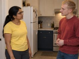 A woman in a yellow shirt and man in a red sweatshirt talking in an apartment kitchen