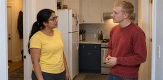 A woman in a yellow shirt and man in a red sweatshirt talking in an apartment kitchen