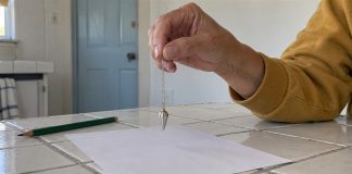 An older adult holding a silver pendulum above blank paper on a white tiled kitchen table.