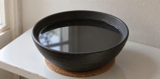 A black ceramic bowl of still water on a cork mat beside a window