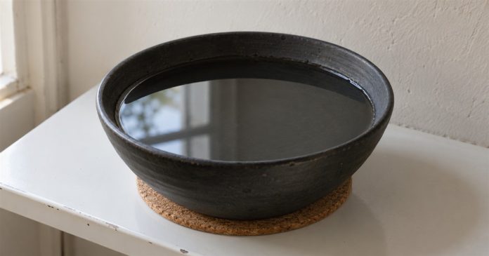 A black ceramic bowl of still water on a cork mat beside a window