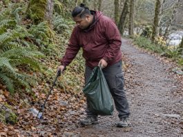 A man in a maroon rain jacket collecting a plastic bottle from a woodland trail