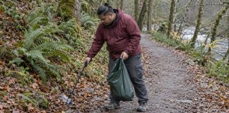 A man in a maroon rain jacket collecting a plastic bottle from a woodland trail