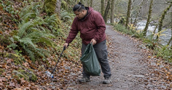 A man in a maroon rain jacket collecting a plastic bottle from a woodland trail