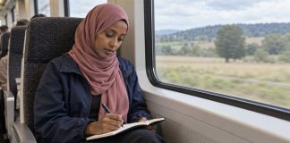 Woman in a dusty-rose headscarf writing in a notebook on her lap beside a train window